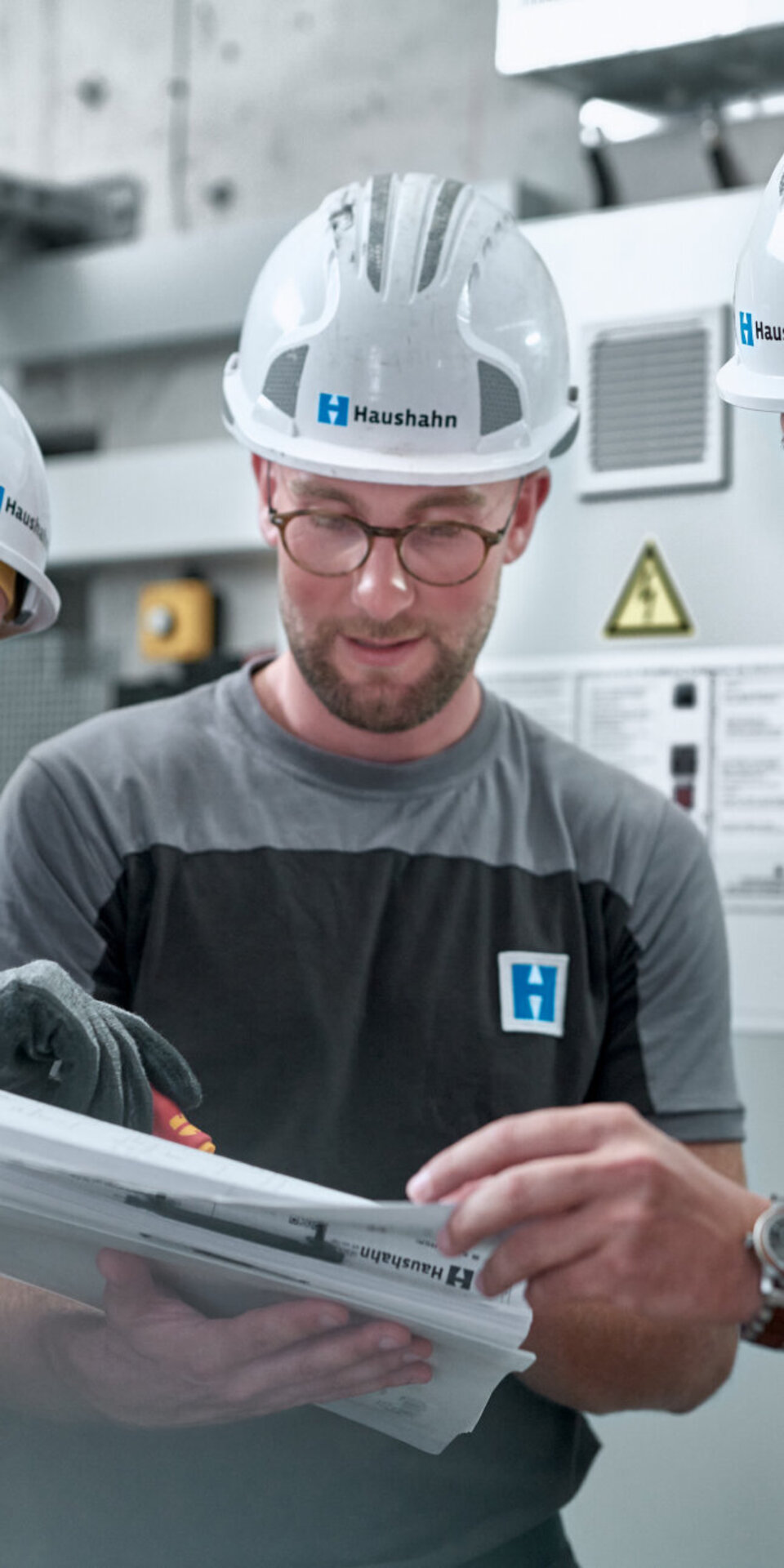 Drei Männer mit weißen Schutzhelmen und Arbeitsuniformen stehen in einem Innenraum zusammen und sehen sich Dokumente oder Pläne genau an. Einer zeigt auf die Papiere, während die anderen aufmerksam zuschauen, was auf Teamwork und Zusammenarbeit schließen lässt.