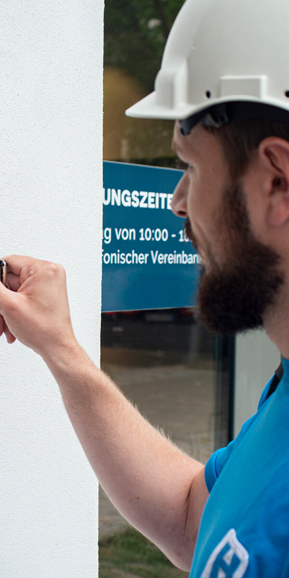 Ein Mann mit weißem Schutzhelm und blauem Hemd betätigt mit einem Werkzeug ein Ventil oder ein Schloss an einer weißen Wand. Im Hintergrund ist eine Glastür mit einem blauen Schild zu sehen.