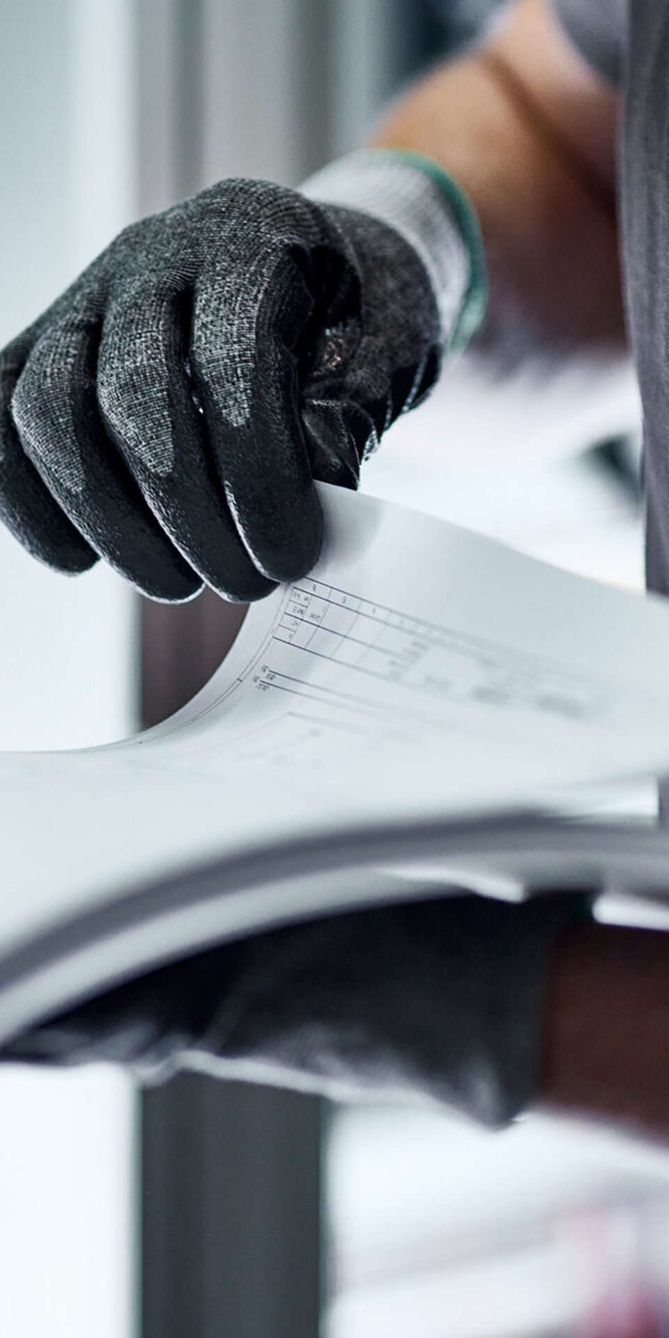 Eine Person mit schwarzen Handschuhen blättert in einem Stapel von Papieren oder einem Dokument, wobei nur ihr Oberkörper und ihre Hände zu sehen sind. Sie trägt ein graues Hemd mit einem blau-weißen Abzeichen oder Logo auf dem Ärmel.