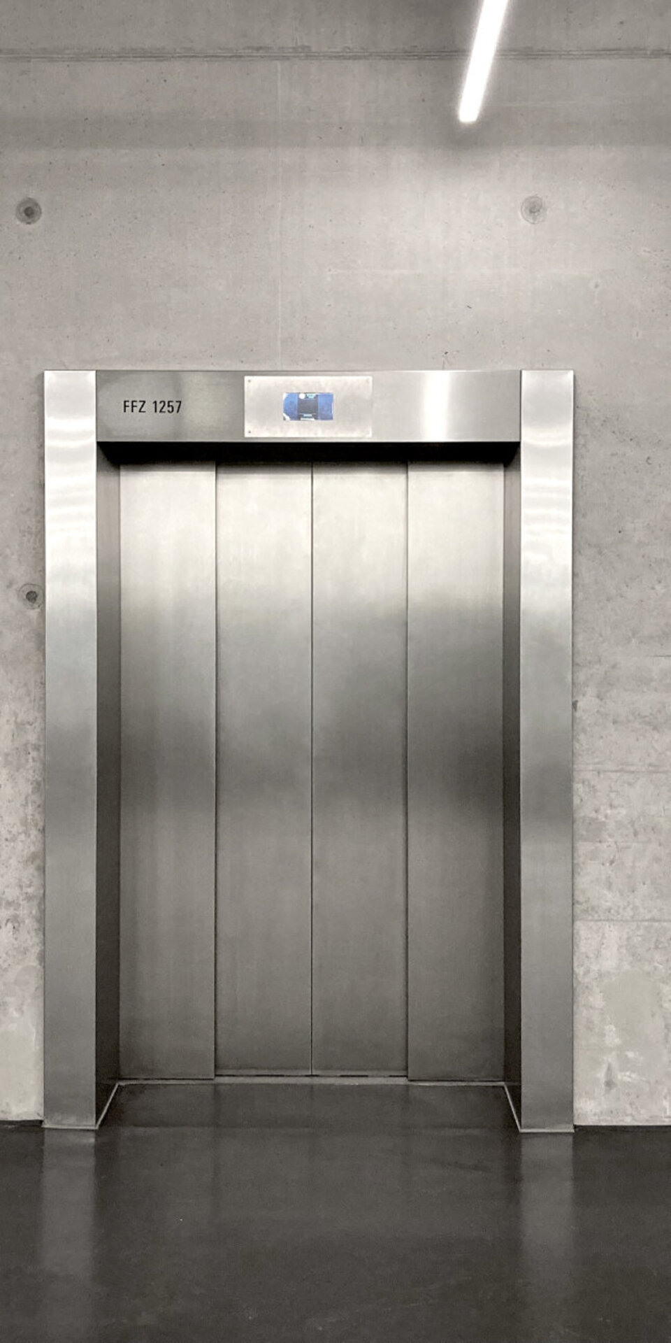 Ein Aufzug aus rostfreiem Stahl mit geschlossenen Türen ist in eine Betonwand eingelassen. In der Nähe sind zwei Aufzugstableaus angebracht. Die Umgebung wirkt industriell und minimalistisch, mit freiliegenden Kabeln und grauen Oberflächen.