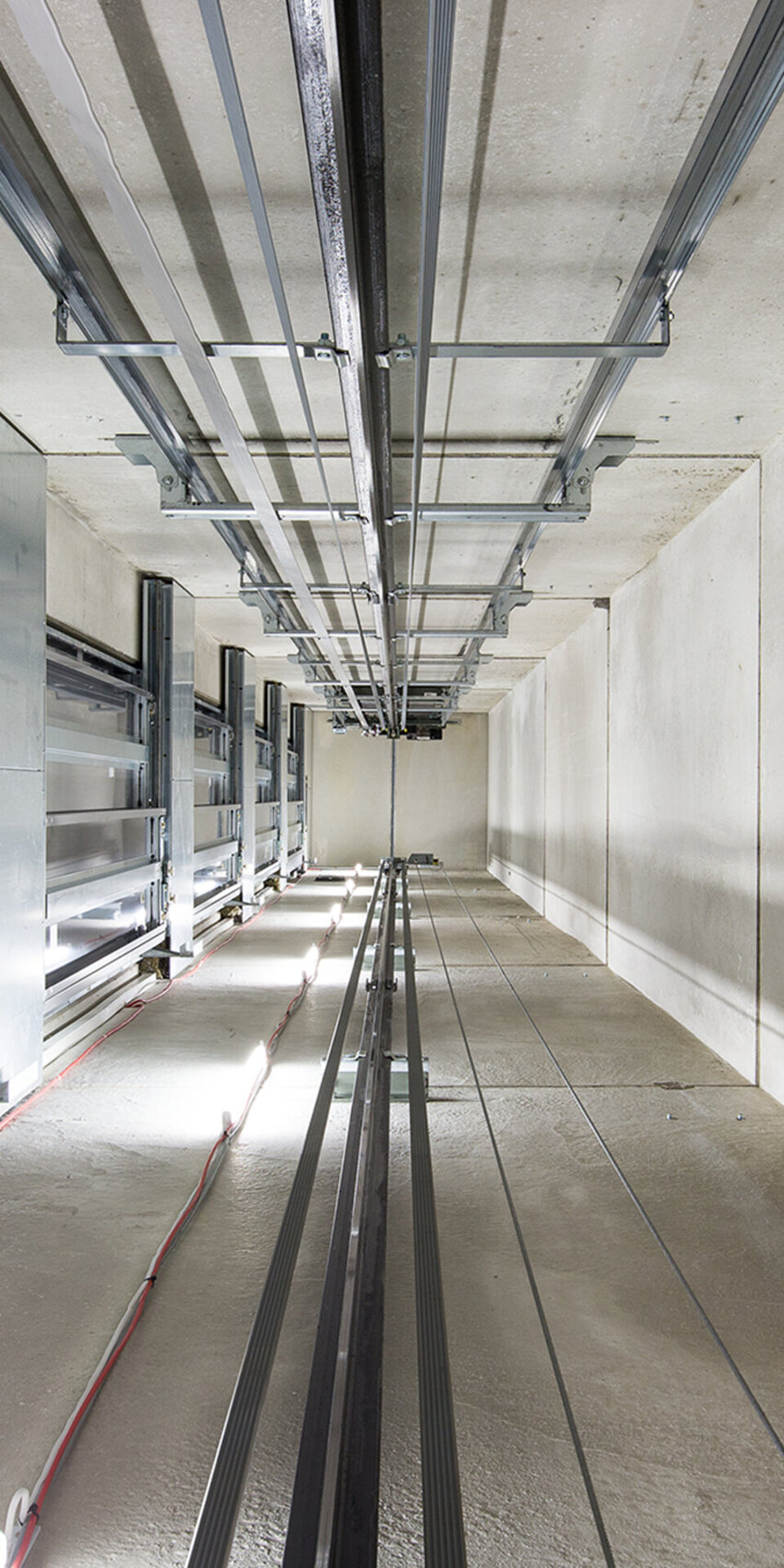 Blick in einen leeren Aufzugsschacht mit sichtbaren Kabeln, Schienen und Rohren, die entlang der Wände und der Decke verlaufen; der Raum ist hell erleuchtet und aus Beton gebaut.