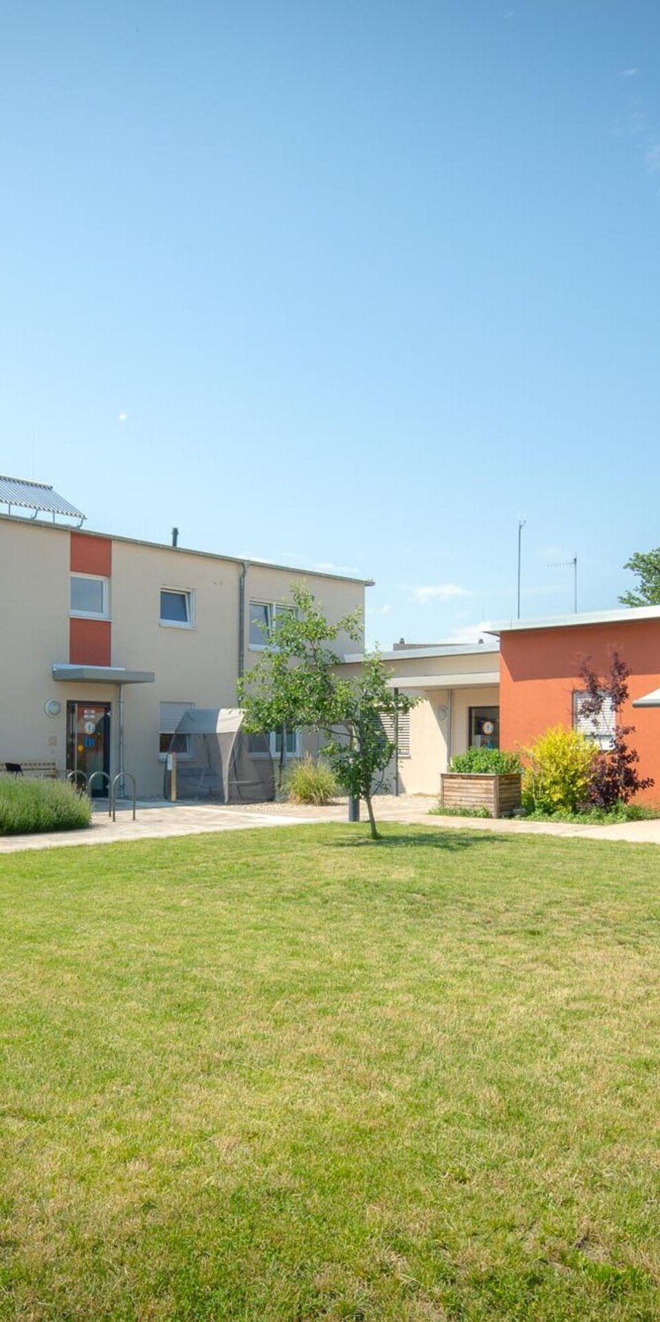 Ein modernes Gebäude mit roten und beigen Wänden steht vor einem gepflegten, grasbewachsenen Innenhof mit einem großen Baum, um den eine runde Holzbank steht, unter einem klaren blauen Himmel.