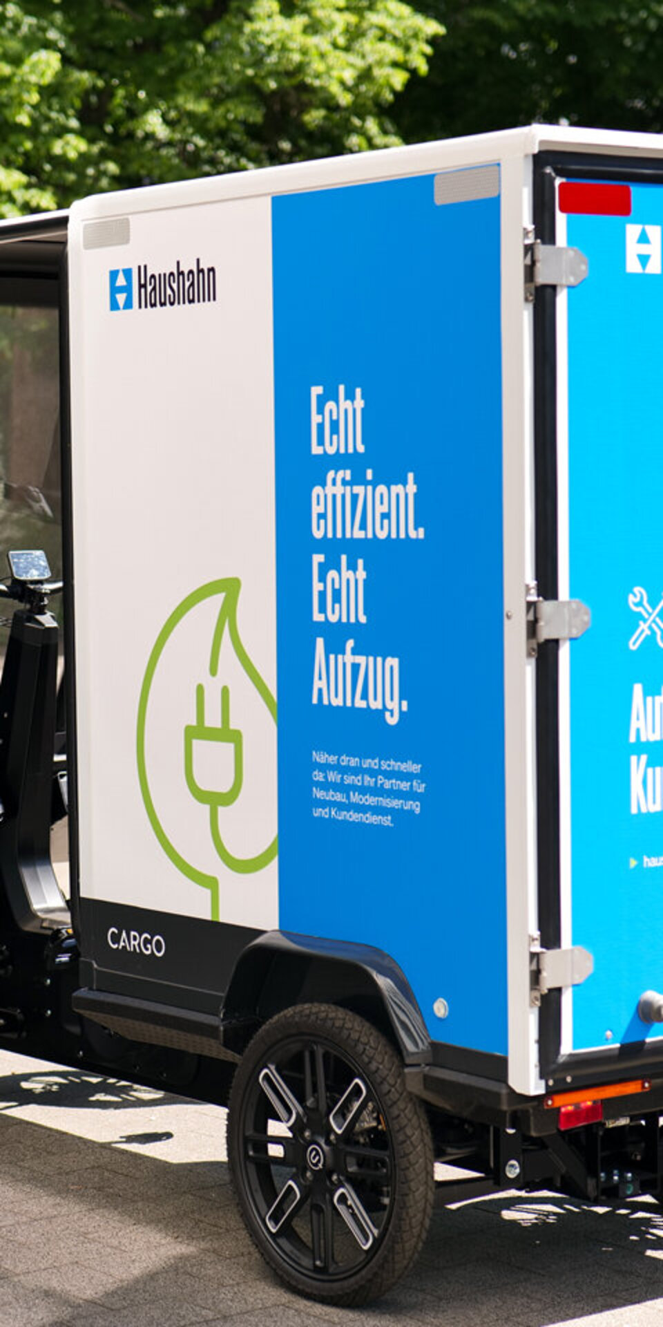 Ein kleiner Elektro-Lastwagen mit Haushahn-Branding ist im Freien geparkt. Auf den Seiten- und Rückseiten ist ein deutscher Text zu sehen, der für den Aufzug und den Kundendienst wirbt, sowie eine Grafik mit einem Stromstecker. Im Hintergrund sind Bäume zu sehen.