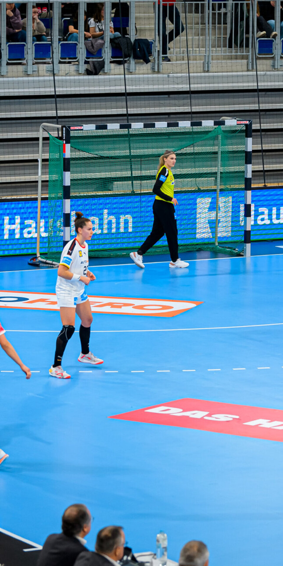 Laufendes Handballspiel der Frauen mit Spielern in weißen und roten Trikots auf einem blauen Spielfeld, einer Torhüterin in der Nähe des Tores und Zuschauern, die im Hintergrund sitzen.