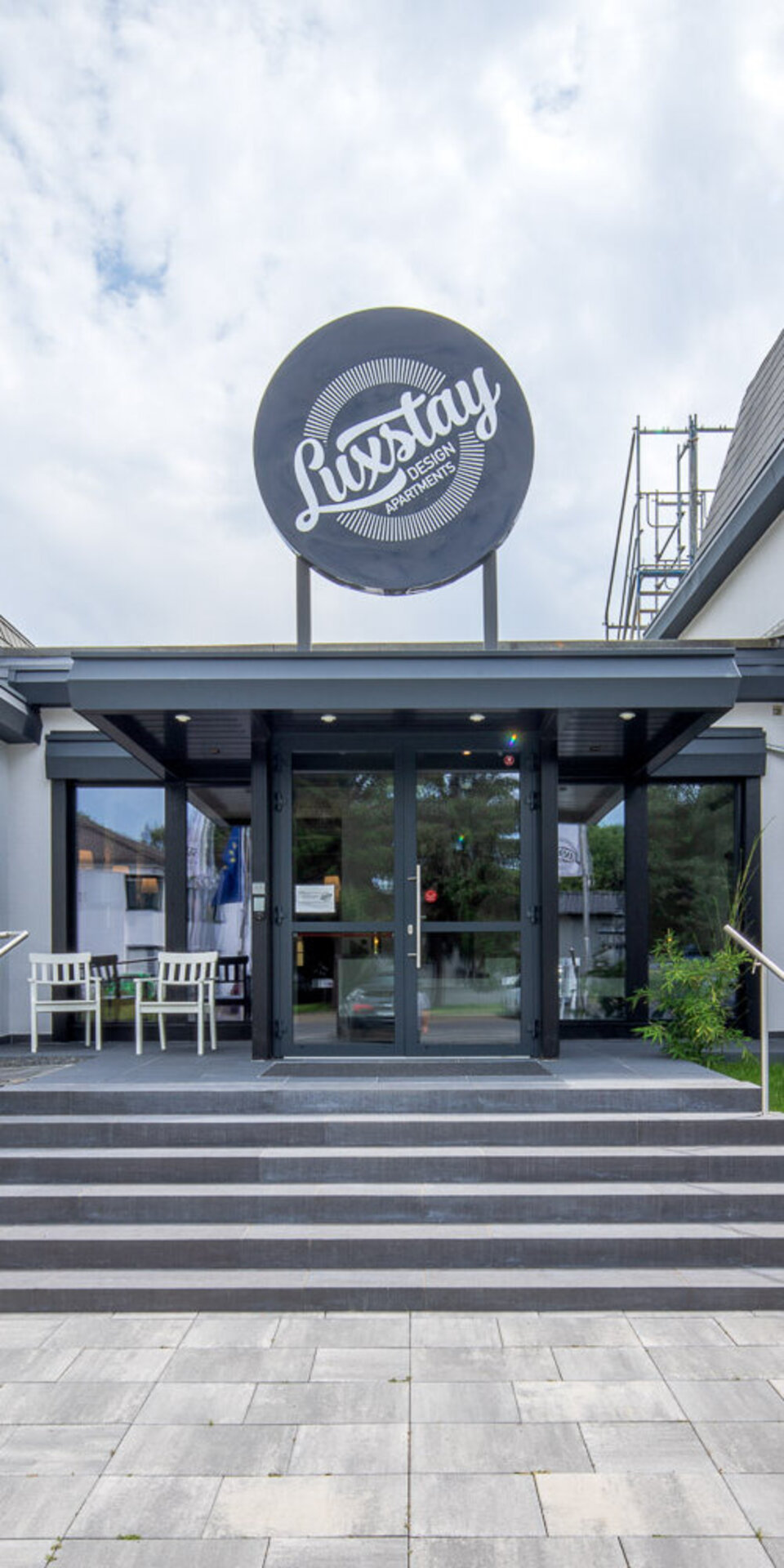 Ein moderner Hoteleingang mit einem großen runden LuxStay-Schild über den Glastüren, grauen Stufen, die nach oben führen, und Grünflächen auf beiden Seiten. Das Gebäude hat weiße Wände und dunkel gerahmte Fenster.