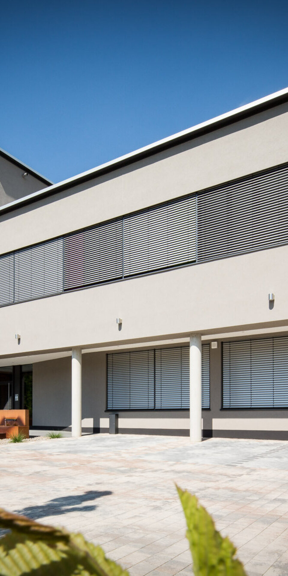 Ein modernes zweistöckiges Gebäude mit großen horizontalen Fenstern und Metallrollos, Flachdach und hellgrauem Äußeren. Der gepflasterte Innenhof ist mit Topfpflanzen bepflanzt, und eine Außentreppe führt in das obere Stockwerk. Darüber blauer Himmel.