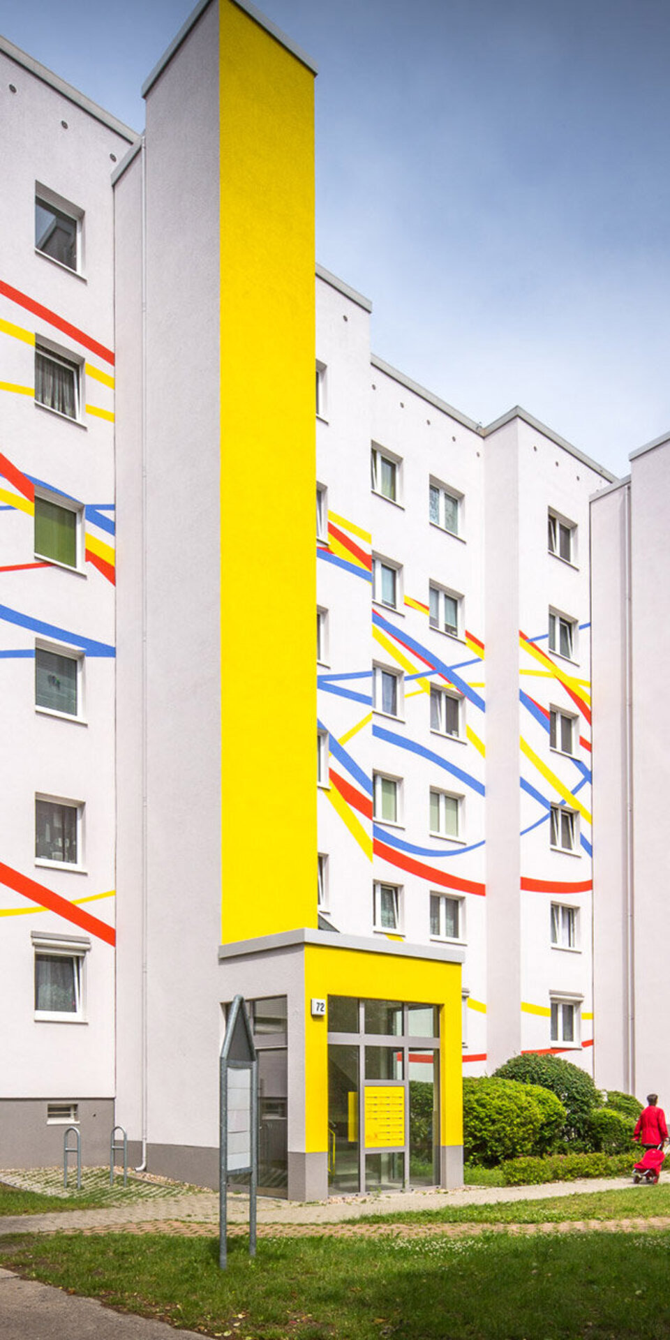 Ein modernes Wohnhaus mit weißen Wänden, verziert mit kräftigen vertikalen Streifen in Gelb, Rot und Blau und bunten Linien, die sich um das Gebäude winden. Bäume und Gras umgeben das Gebäude an einem hellen Tag.