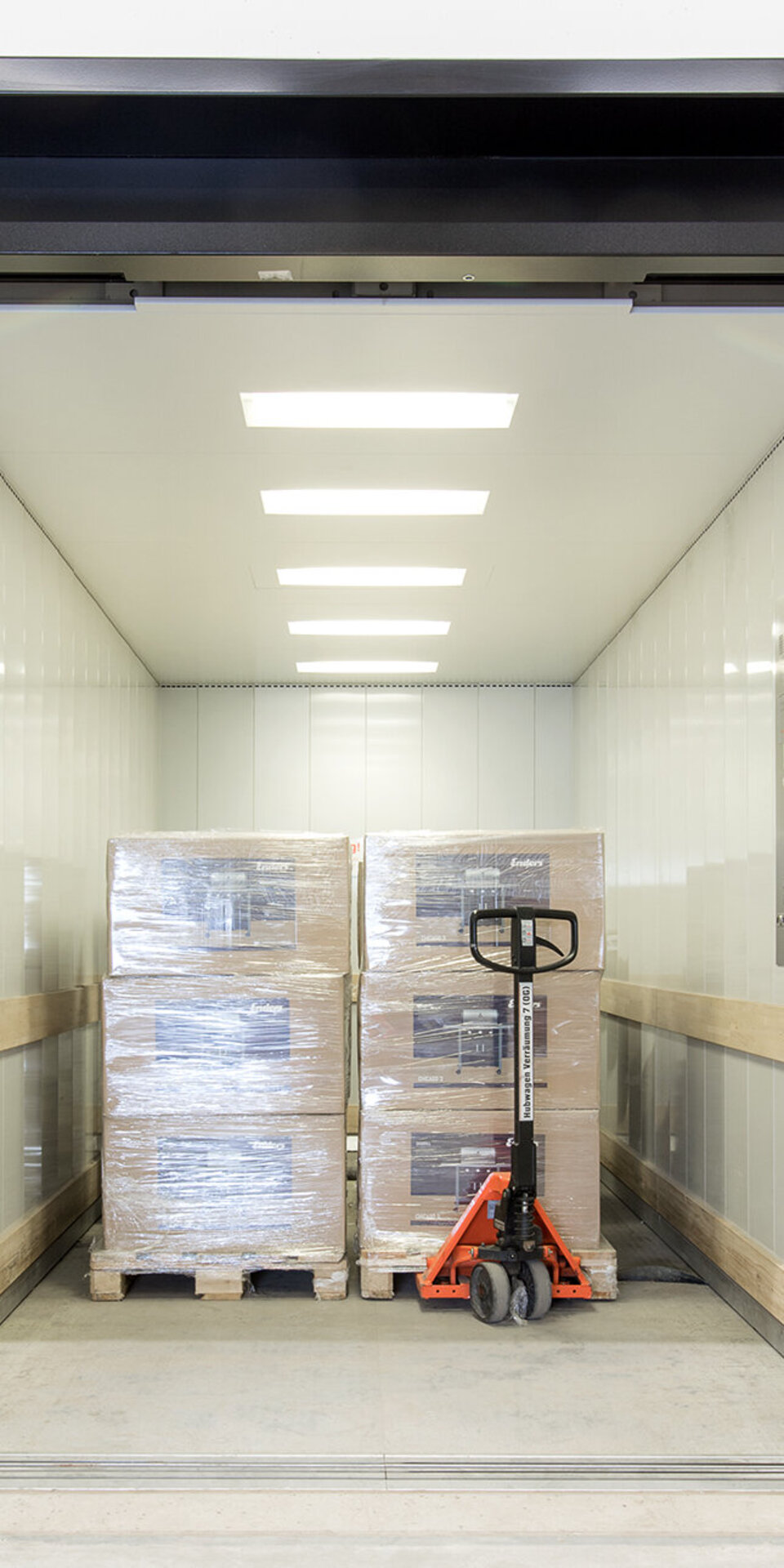 Ein großer Lastenaufzug mit heller Deckenbeleuchtung enthält zwei gestapelte Paletten mit verpackten Kisten und einen orangefarbenen Hubwagen. Der Aufzug hat Schutzschienen an den Wänden und gelbe Sicherheitspoller am Eingang.