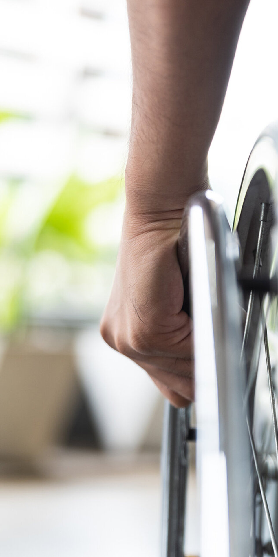 Nahaufnahme der Hand einer Person, die das Rad eines Rollstuhls festhält, mit einem unscharfen Hintergrund mit grünen Pflanzen und natürlichem Licht.