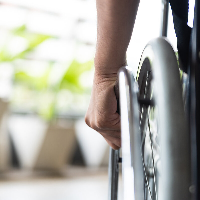 Nahaufnahme der Hand einer Person, die das Rad eines Rollstuhls festhält, mit einem unscharfen Hintergrund mit grünen Pflanzen und natürlichem Licht.