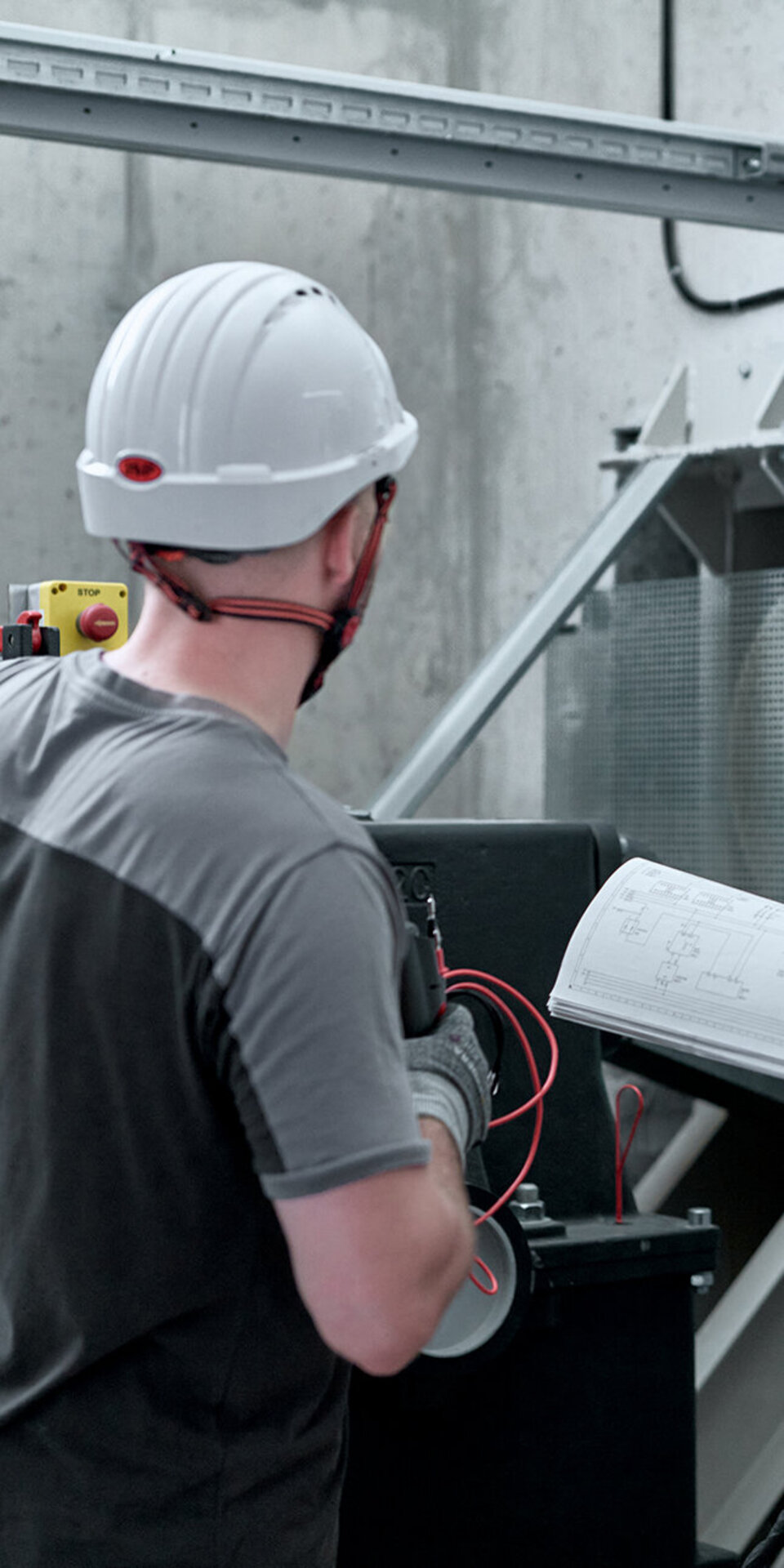 Zwei Arbeiter mit Schutzhelmen und Sicherheitsausrüstung überprüfen Maschinen in einer industriellen Umgebung. Der eine zeigt auf die Ausrüstung, während der andere ein technisches Handbuch oder eine Blaupause aufschlägt, beide konzentriert auf ihre Aufgabe.