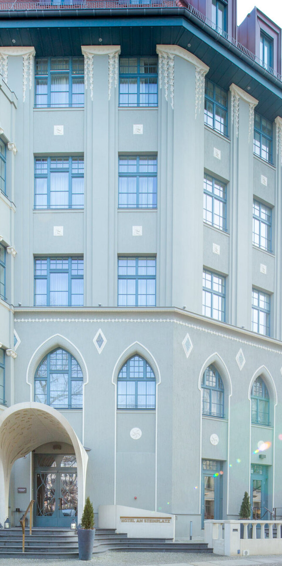 Elegantes, hellgraues Hotel mit gewölbten Fenstern, dekorativen Balkonen und einem verschnörkelten Eingang. Das Gebäude weist geometrische und klassische architektonische Elemente auf, mit Topfpflanzen in der Nähe der Eingangstür und Sonnenlicht, das die Fassade erhellt.