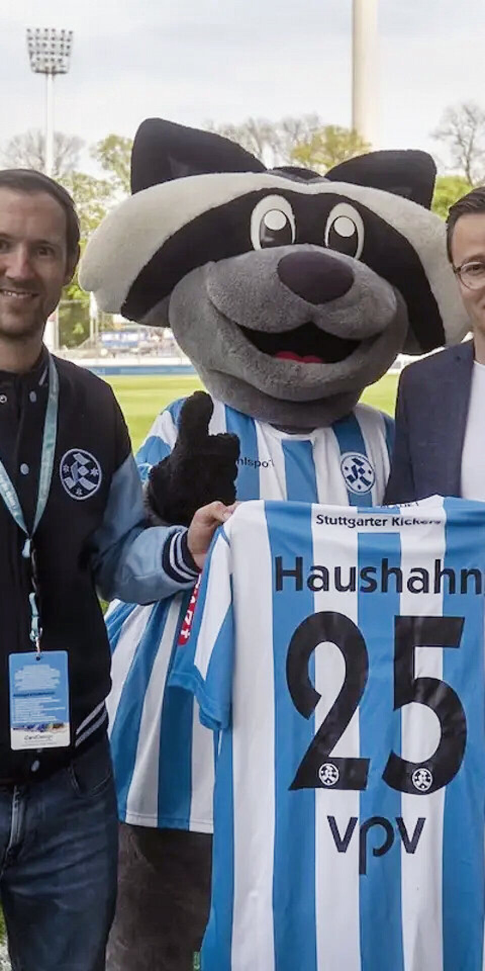 Zwei Männer stehen lächelnd auf einem Fußballfeld mit einem Waschbär-Maskottchen. Das Maskottchen trägt ein blau-weiß gestreiftes Trikot mit der Aufschrift Haushahn 25. Im Hintergrund sind Stadionplätze und Bäume zu sehen.