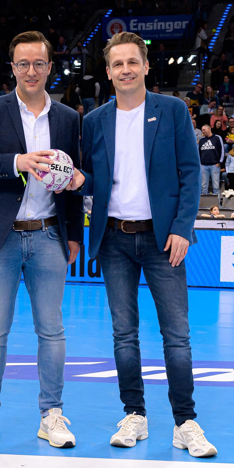 Zwei Männer in Blazern, Jeans und weißen Turnschuhen stehen auf einem Hallensportplatz, einer hält einen Handball. Die Zuschauer füllen die Plätze hinter ihnen. Die Atmosphäre ist hell und lebendig.