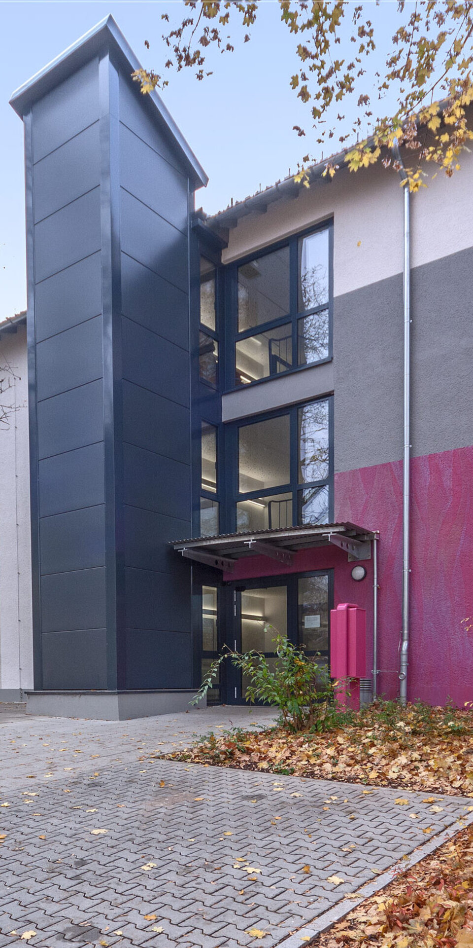 Ein modernes, zweistöckiges Gebäude mit grauen und kastanienbraunen Akzenten, großen Fenstern und einem zentralen Glaseingang. Herbstliche Bäume und goldenes Laub säumen den gepflasterten Weg, der zum Eingang führt.