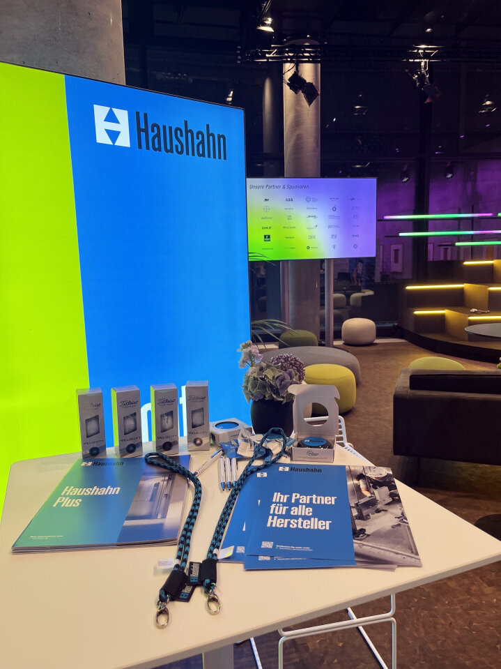 Ein Tischaufsteller mit blauen und grünen Haushahn-Broschüren, Schlüsselanhängern, Schlüsselbändern und kleinen Gadgets. Im Hintergrund leuchtet ein großer Bildschirm mit dem Haushahn-Logo, Lounge-Sitzgelegenheiten und Neonlichter sind in einem modernen Innenbereich zu sehen.