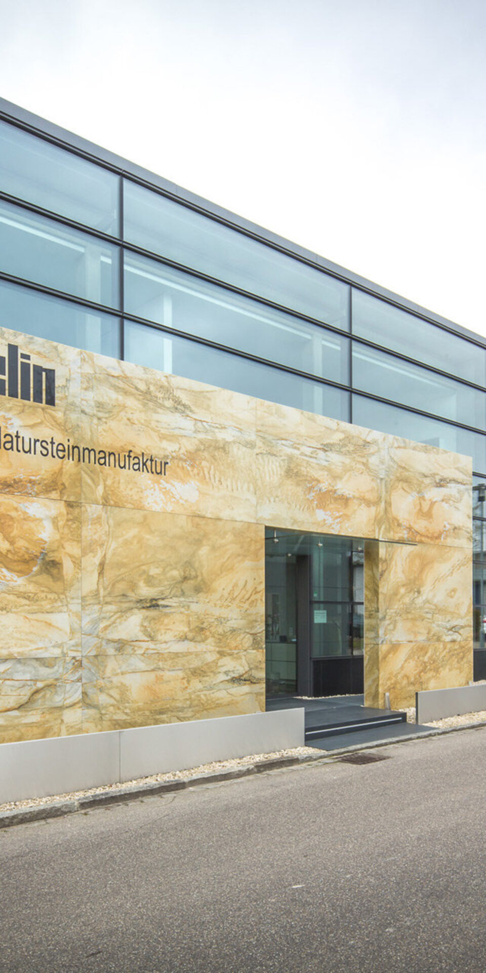 Modernes Gebäude mit großen Glasfenstern, einem hellbraunen Steineingang mit der Aufschrift Stärchelin Die exklusive Natursteinmanufaktur, weißen Außenverkleidungen und einer Reihe ordentlich gestutzter Bäume entlang des Gehwegs.