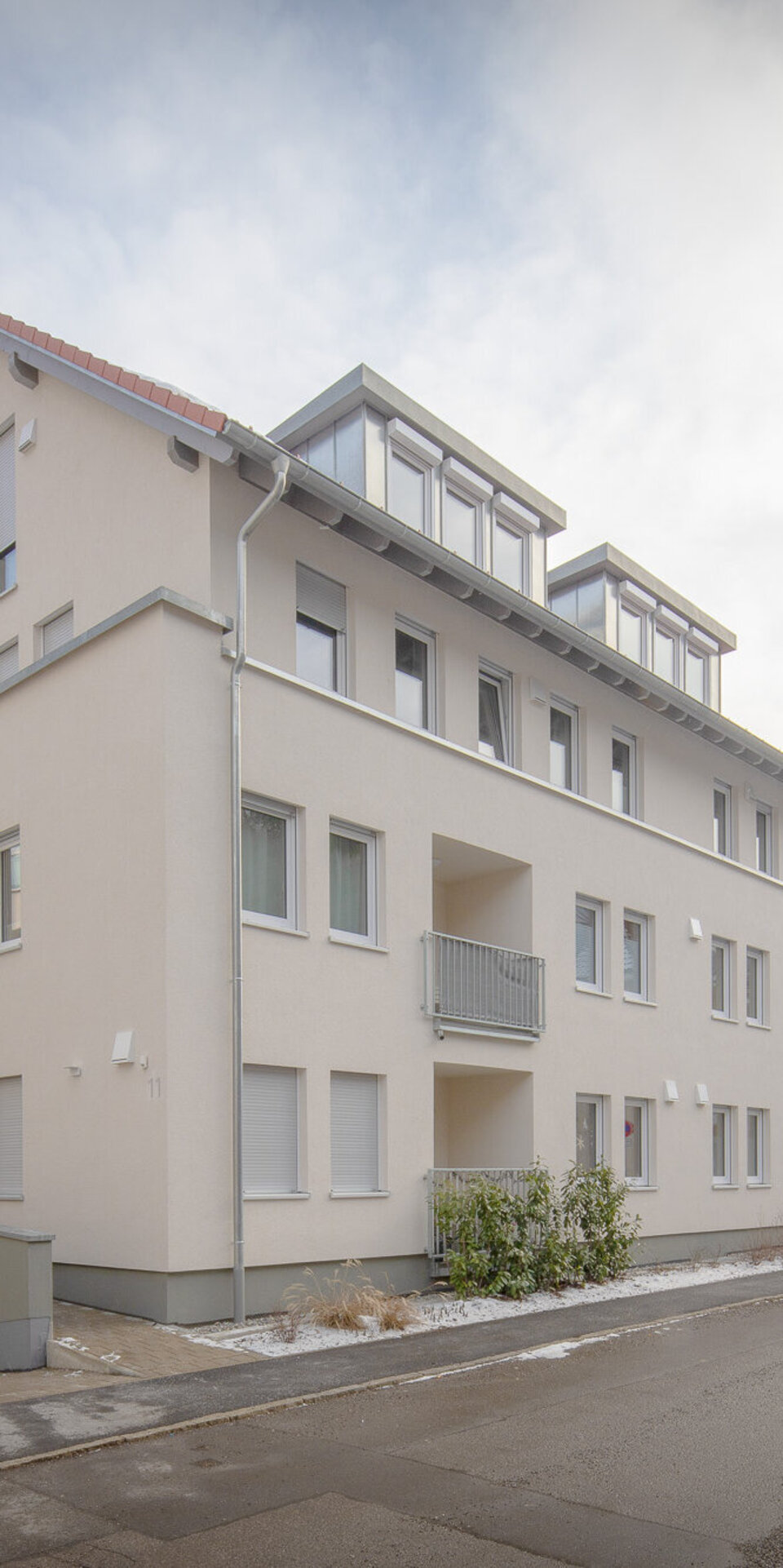 Ein modernes, dreistöckiges, beigefarbenes Mehrfamilienhaus mit vielen Fenstern und Balkonen steht an einer ruhigen, verschneiten Straße. Entlang der Straße sind Autos geparkt, und auf der linken Seite ist die Einfahrt eines Parkhauses zu sehen.