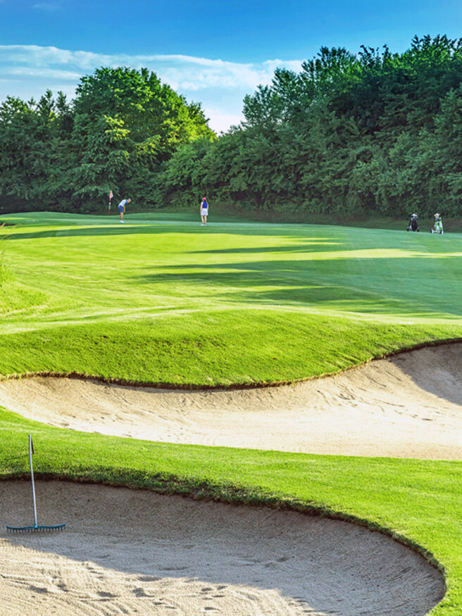 Ein grüner Golfplatz mit Sandbunkern im Vordergrund, einer Harke, die auf dem Sand ruht, und mehreren Golfspielern, die in der Ferne unter blauem Himmel spazieren gehen und spielen, während im Hintergrund Bäume stehen.