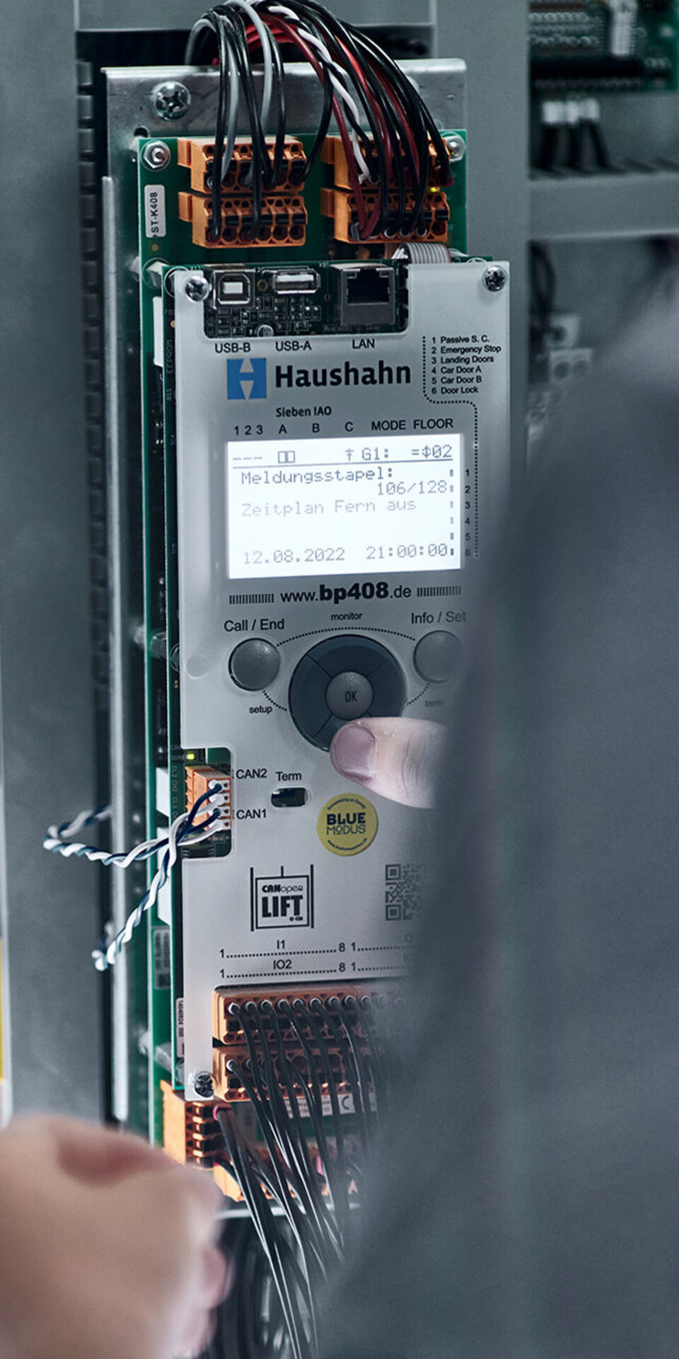 Eine Person interagiert mit einer Hausbahn-Aufzugstafel in einem Schaltschrank mit sichtbaren Kabeln und einem beleuchteten Display, das technische Informationen anzeigt.