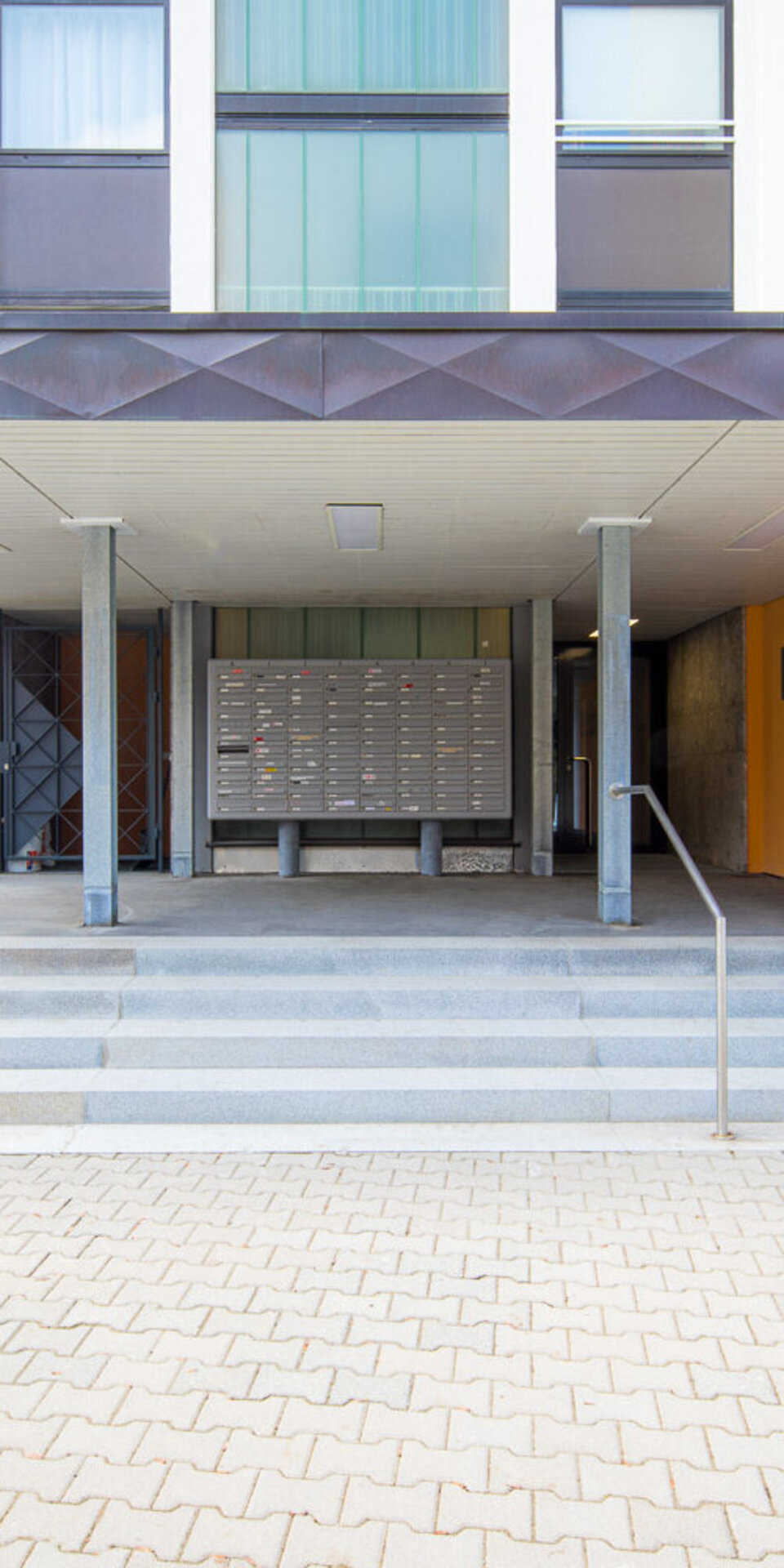 Eingang zu einem modernen Wohnhaus mit breiten Stufen, einer Rampe mit Geländer, mehreren Briefkästen und der Adresse Leebstr. 9 an einer gelben Wand. Auf der linken Seite befinden sich Büsche und ein roter Briefkasten.