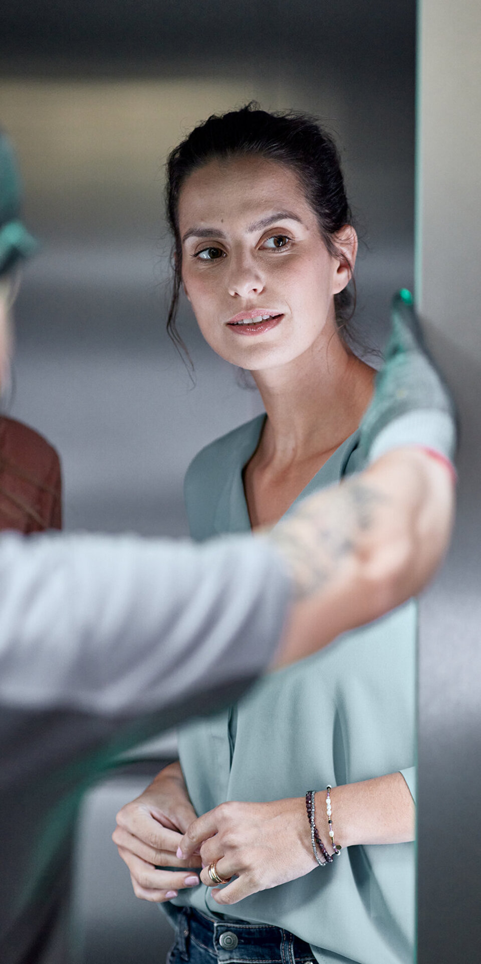 Eine Frau in hellblauer Bluse steht in einem Haus und schaut aufmerksam auf eine Person im Vordergrund, die gestikuliert und Arbeitshandschuhe und eine Mütze trägt. Die Umgebung wirkt professionell oder industriell.