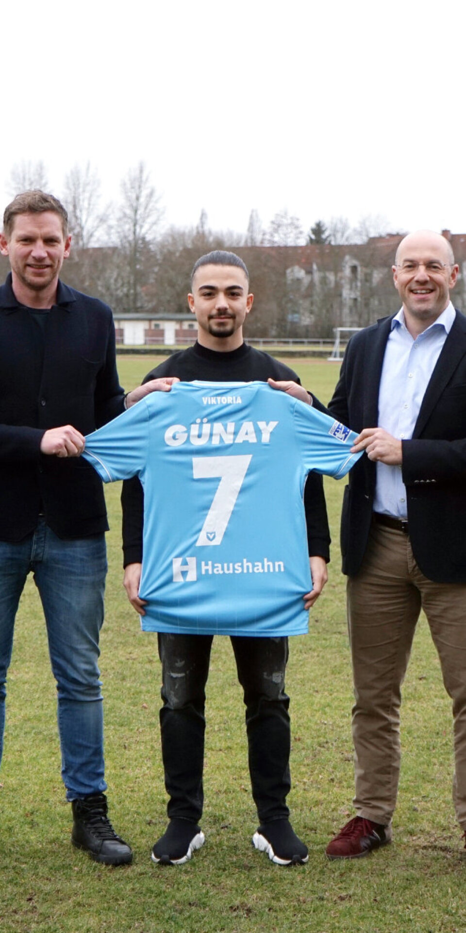 Fünf Männer stehen auf einem Fußballfeld, wobei zwei Männer in der Mitte ein blaues Fußballtrikot mit der Aufschrift GÜNAY 7 tragen. Drei von ihnen tragen Fußballkleidung, zwei sind in Business-Kleidung gekleidet. Im Hintergrund sind Bäume und Gebäude zu sehen.