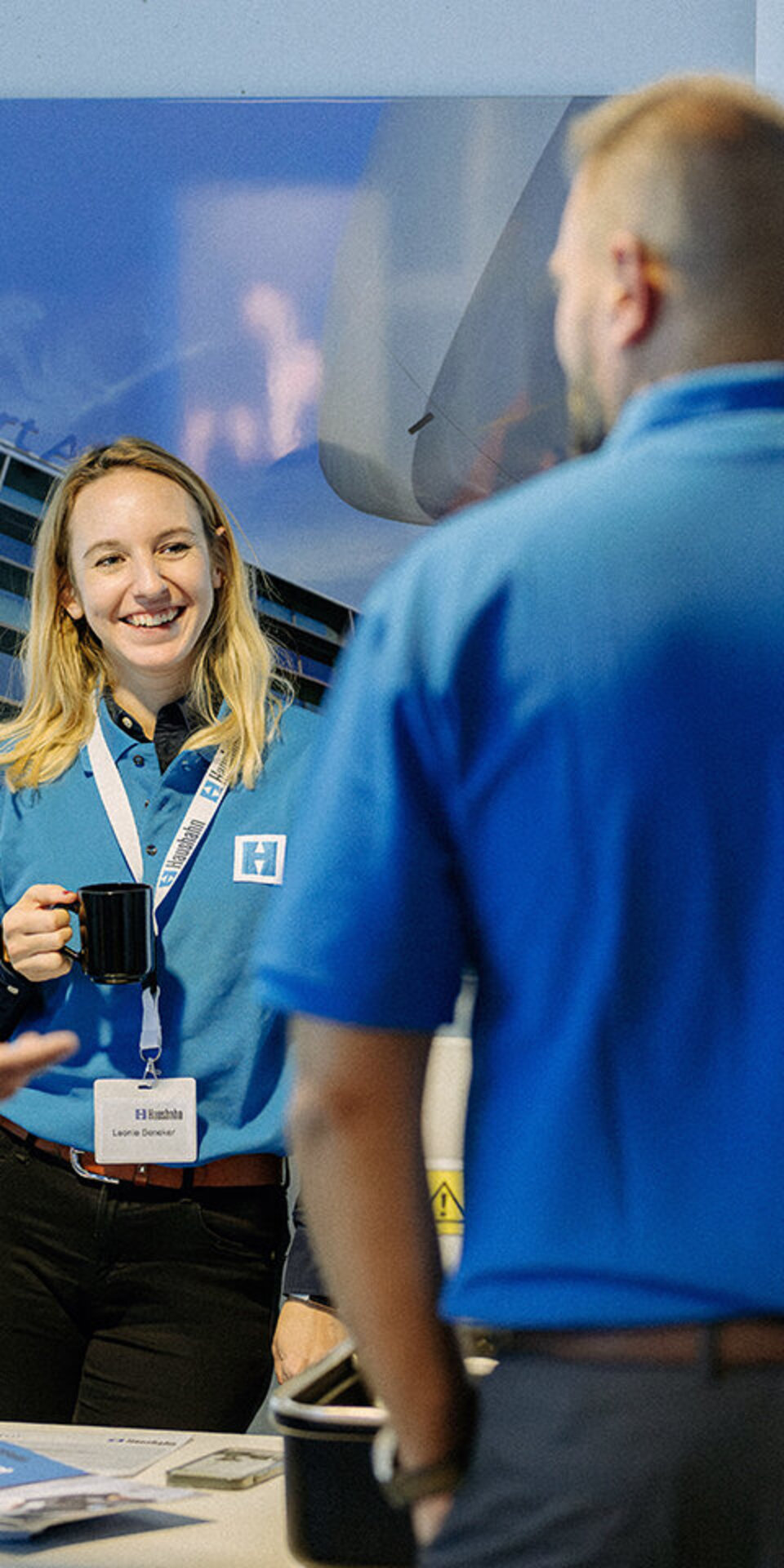 Vier Personen, drei Männer und eine Frau, unterhalten sich und lächeln bei einer Veranstaltung in einer Halle. Die Frau hält eine schwarze Tasse in der Hand und trägt ein Namensschild; zwei Männer tragen passende blaue Hemden. Hinter ihnen hängt ein großes Foto an der Wand.