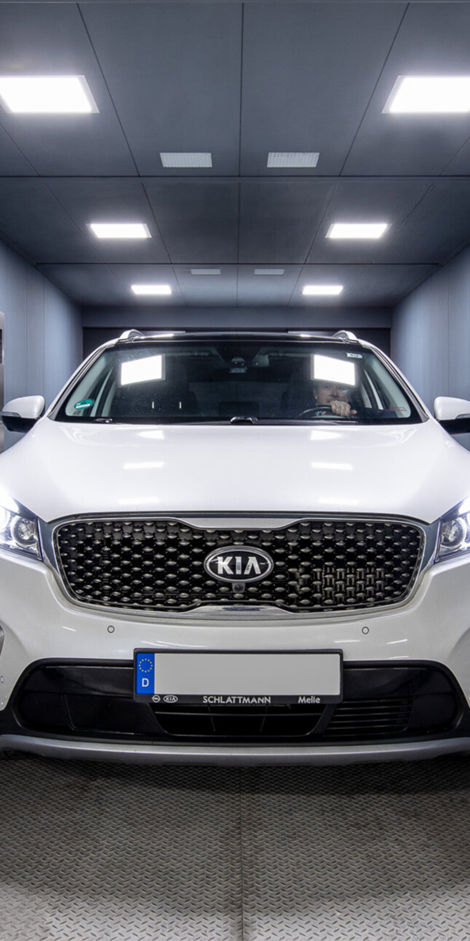 Ein weißer Kia-Geländewagen ist in einem gut beleuchteten, geschlossenen Raum mit grauen Wänden und Deckenleuchten geparkt und blickt direkt auf die Kamera. Die Scheinwerfer des Fahrzeugs sind eingeschaltet.