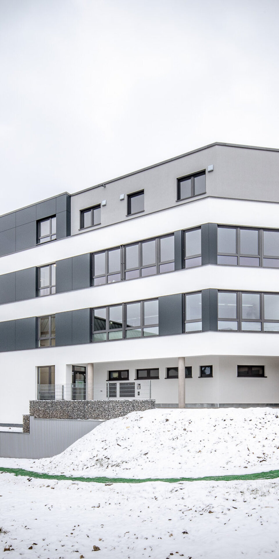 Ein modernes, mehrstöckiges weißes Gebäude mit großen Fenstern, Flachdach und grauen Akzenten steht auf einem verschneiten Hügel neben anderen Wohngebäuden unter einem bewölkten Himmel.