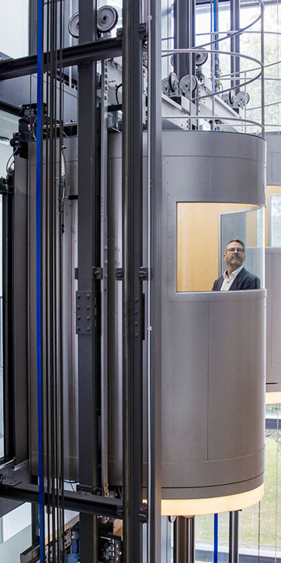 Ein Mann im Anzug steht in einem modernen, zylindrischen Glasaufzug, der in einem hellen Gebäude mit großen Fenstern und mehreren Aufzugsschächten nach oben fährt.