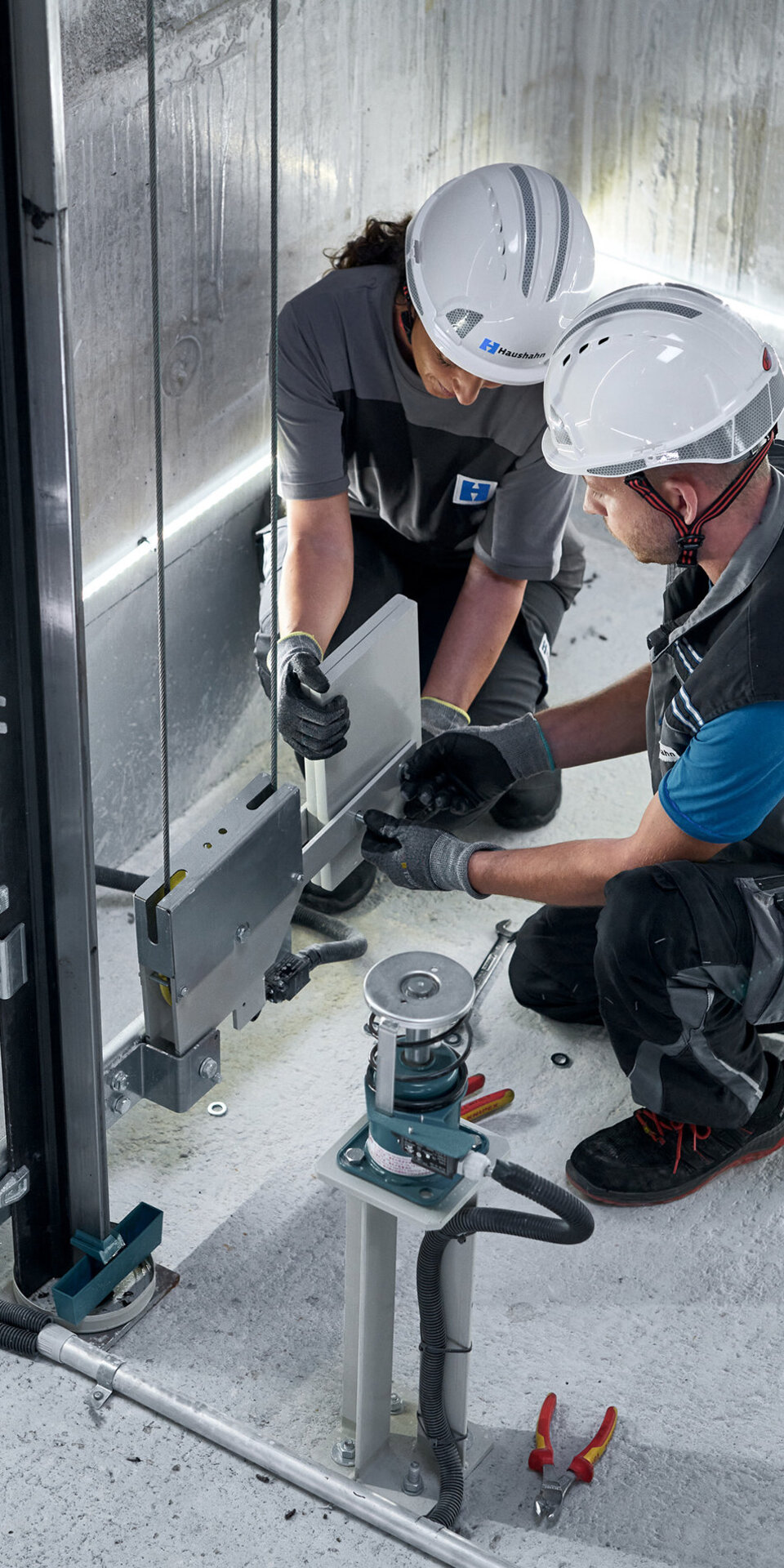 Zwei Arbeiter mit Helmen und Schutzausrüstung knien auf einem Betonboden und installieren oder reparieren mechanische Geräte in einer gut beleuchteten Industrie- oder Bauumgebung. Um sie herum sind Werkzeuge und Kabel zu sehen.