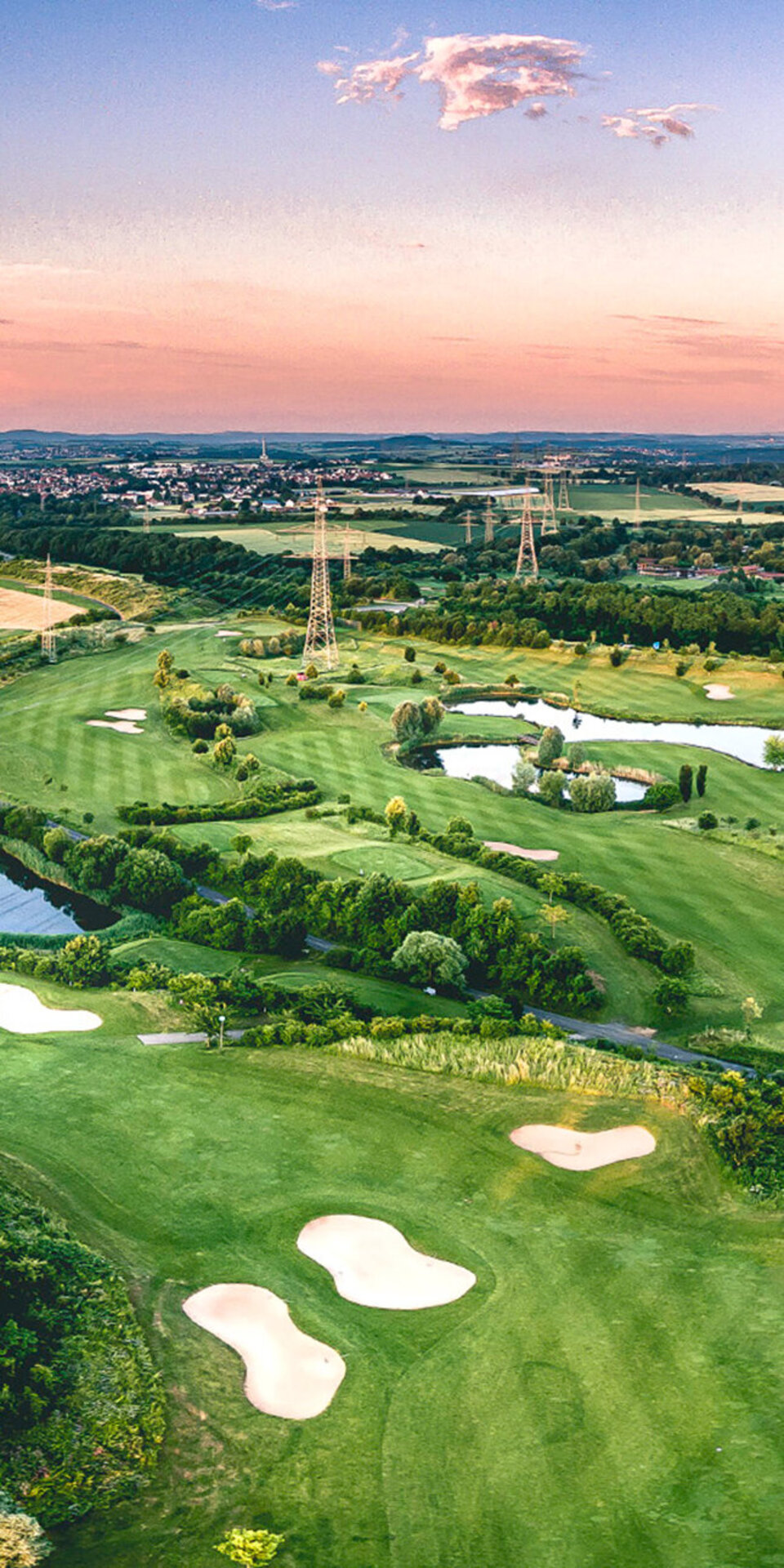 Luftaufnahme eines üppig grünen Golfplatzes mit Sandbunkern, Teichen und Baumgruppen vor einer hügeligen Landschaft und einer entfernten Stadt unter einem farbenfrohen Sonnenuntergangshimmel.