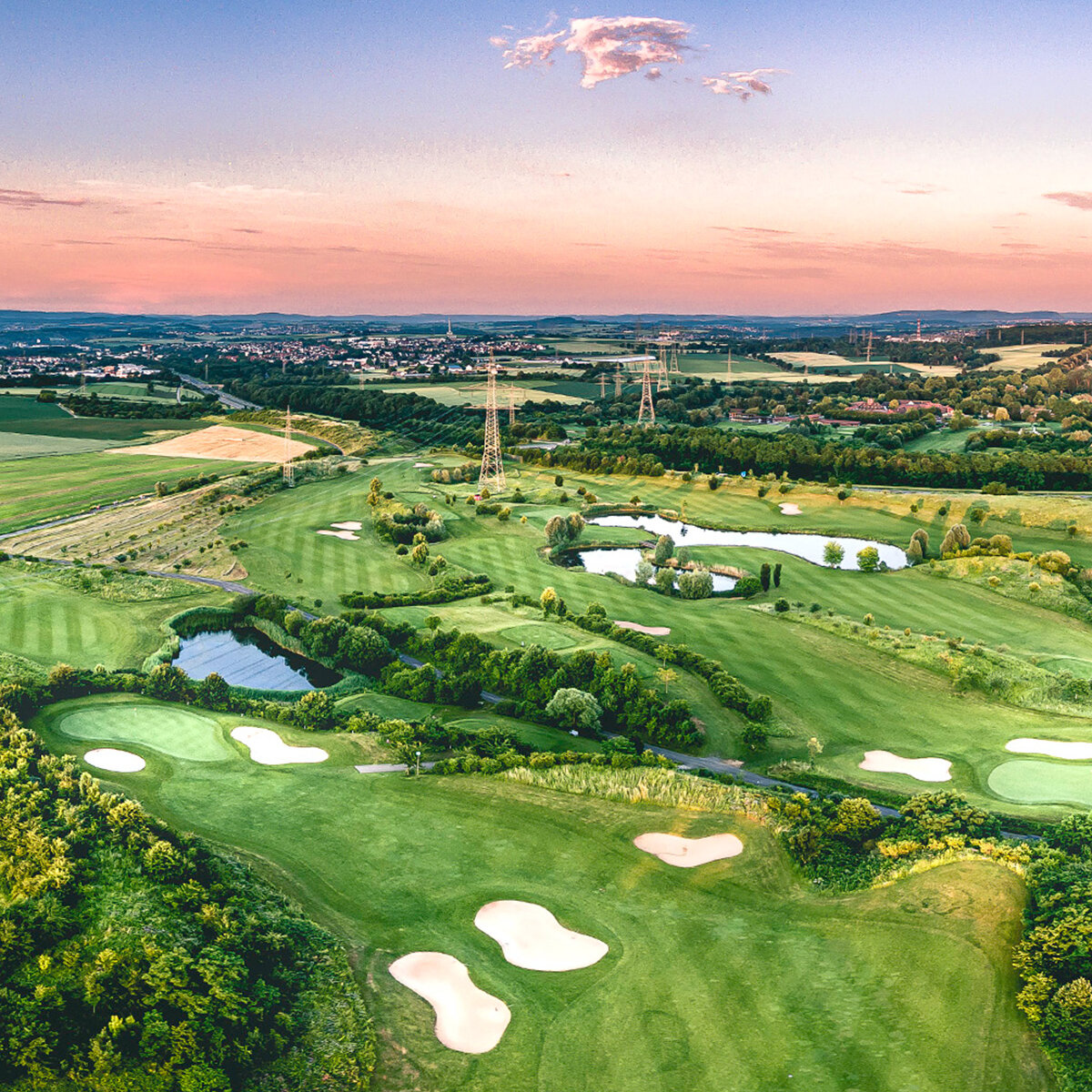 Luftaufnahme eines üppig grünen Golfplatzes mit Sandbunkern, Teichen und Baumgruppen vor einer hügeligen Landschaft und einer entfernten Stadt unter einem farbenfrohen Sonnenuntergangshimmel.