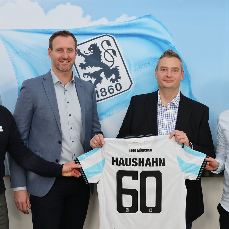 Vier Männer stehen vor einer Wand mit einer großen Fahne mit dem Logo von 1860 München. Zwei von ihnen halten ein Fußballtrikot mit Haushahn und der Nummer 60 auf dem Rücken.