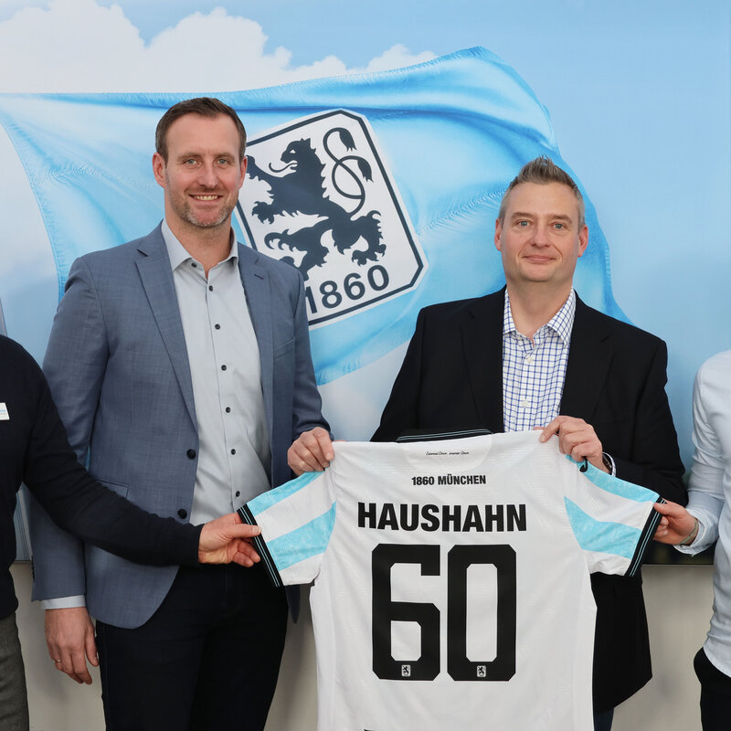 Vier Männer stehen vor einer Wand mit einer großen Fahne mit dem Logo von 1860 München. Zwei von ihnen halten ein Fußballtrikot mit Haushahn und der Nummer 60 auf dem Rücken.