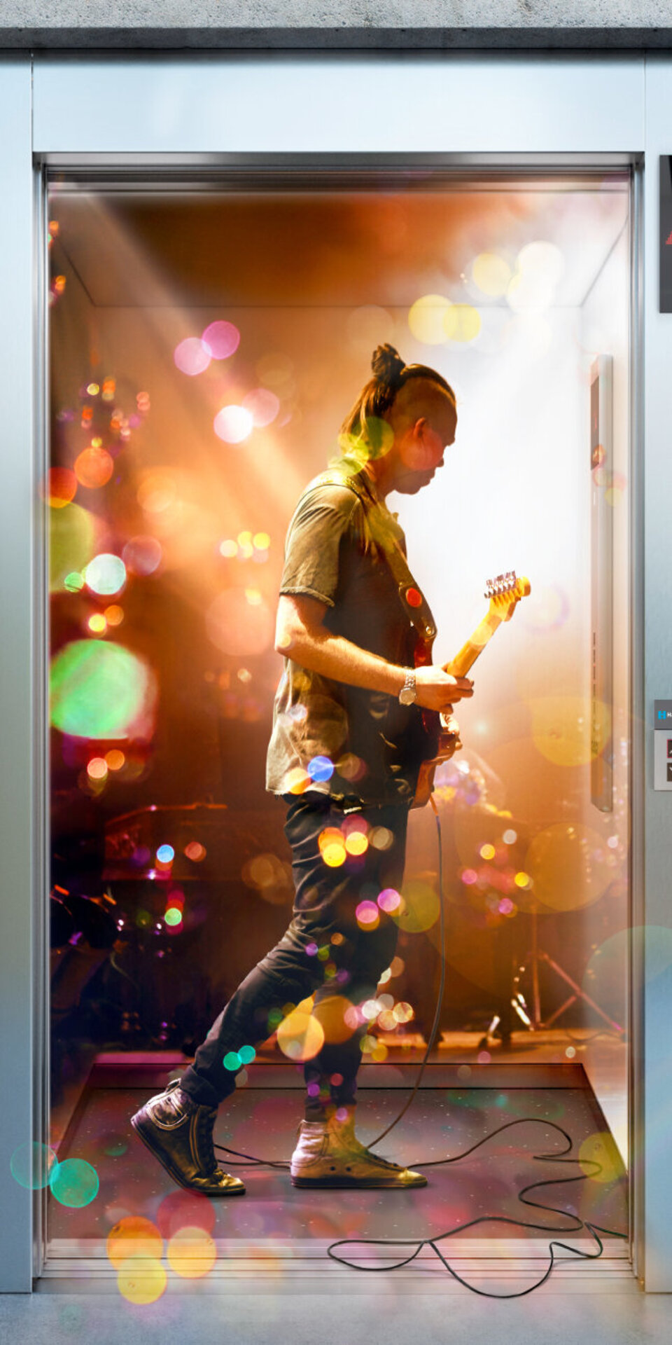 Eine E-Gitarre spielende Person steht in einem hell erleuchteten Aufzug, umgeben von bunten Lichteffekten und Reflexionen, die eine lebendige und surreale Atmosphäre schaffen.