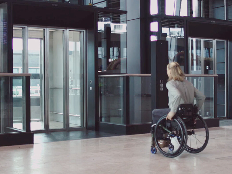 Eine Frau im Rollstuhl nähert sich einem gläsernen Aufzug in einem modernen Gebäude mit großen Fenstern und schlichten, schwarzen Strukturelementen.