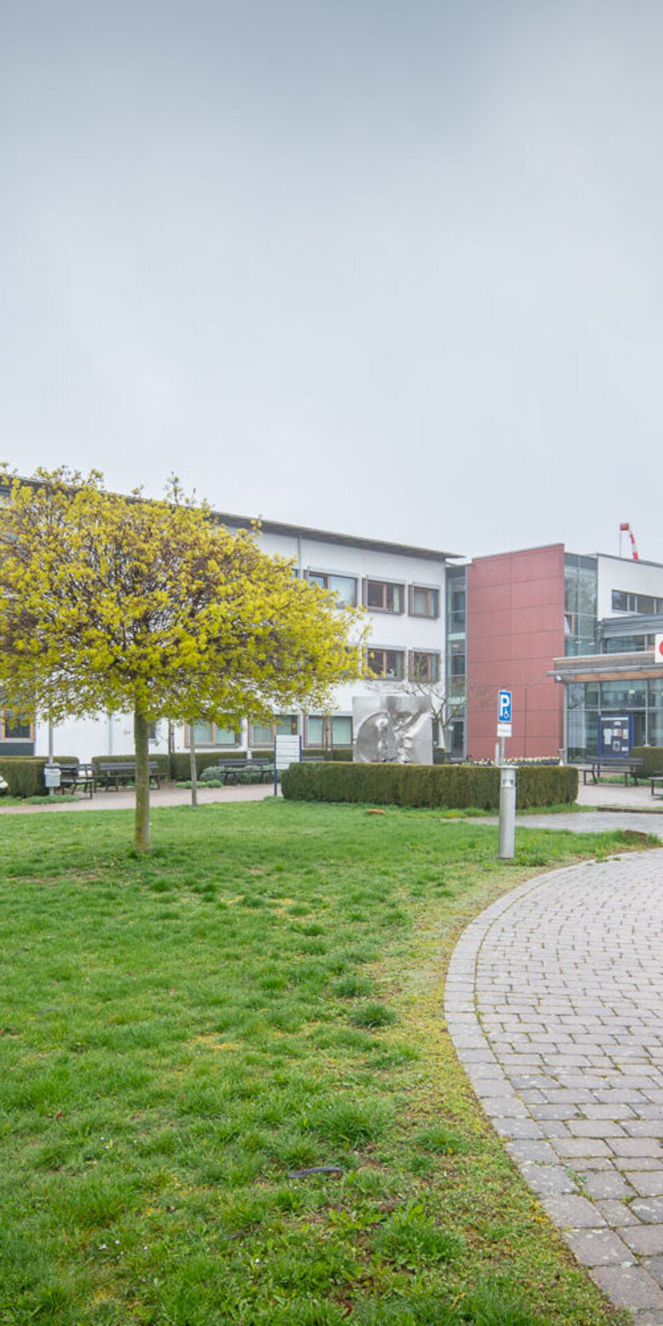 Ein modernes Gebäude mit großen Fenstern und einer roten Eingangsfassade, umgeben von grünem Gras, Bäumen und einem geschwungenen Steinweg an einem bewölkten Tag.