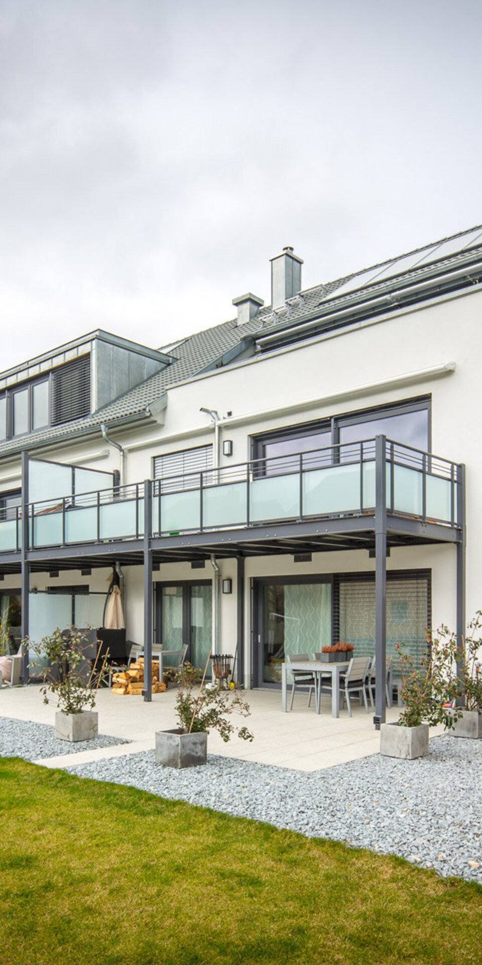 Modernes zweistöckiges weißes Haus mit großen Fenstern, Balkonen und einem geräumigen Terrassenbereich. Das Haus hat ein Schrägdach, einen gepflegten Rasen und befindet sich in einem Vorort mit geparkten Autos und bewölktem Himmel.