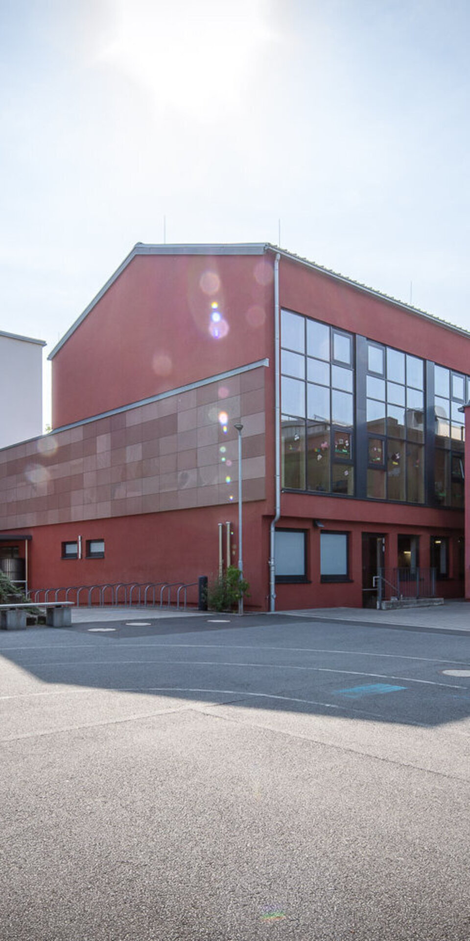 Ein modernes Schulgebäude mit großen Fenstern und einer roten Fassade steht neben einem weißen mehrstöckigen Gebäude. Im Vordergrund befinden sich ein leerer Innenhof und eine Treppe unter einem klaren, sonnigen Himmel.