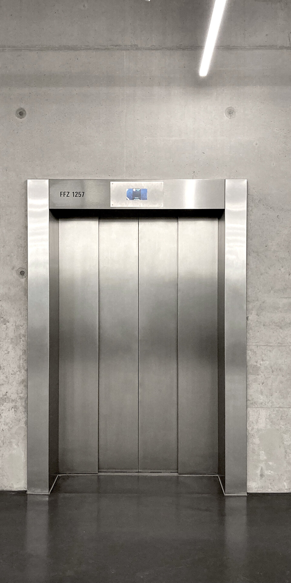 Drei geschlossene Aufzugstüren aus rostfreiem Stahl sind in eine graue Betonwand eingelassen, neben denen sich jeweils ein Bedienfeld befindet. Darüber ist eine einzige Leuchtstoffröhre zu sehen, und der Boden ist dunkel.