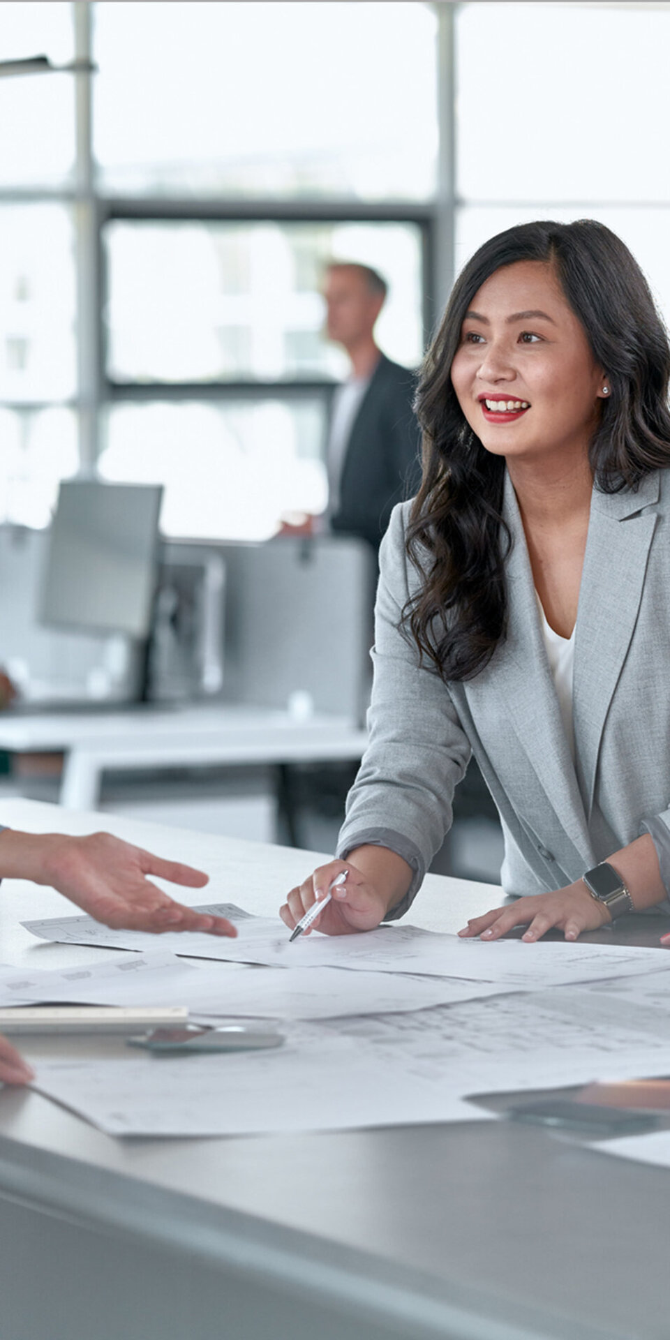 Vier Fachleute versammeln sich um einen mit Dokumenten bedeckten Tisch und führen eine Diskussion. Eine Frau im grauen Blazer lächelt und zeigt auf die Papiere, was auf eine Zusammenarbeit in einer modernen Büroumgebung hindeutet.