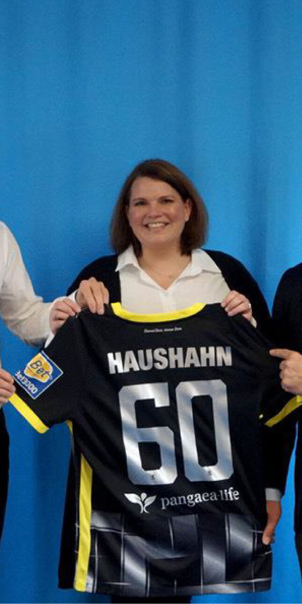 Drei Personen lächeln und halten ein schwarz-gelbes Sporttrikot mit der Aufschrift HAUSHAHN 60 hoch, das vor einem blauen Hintergrund mit einem Löwenwappen und der Nummer 1860 in der oberen rechten Ecke steht.