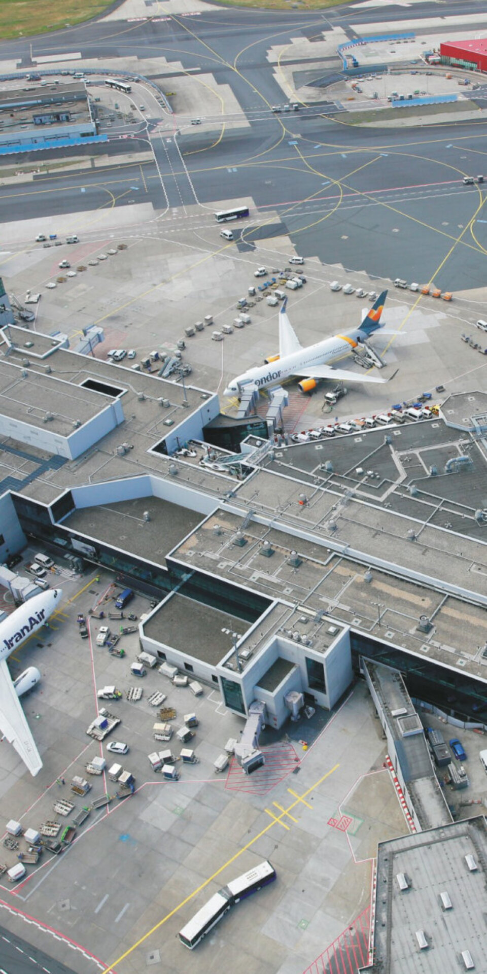 Luftaufnahme eines belebten Flughafenterminals mit mehreren Flugzeugen an den Gates, Fluggastbrücken, Servicefahrzeugen und Start- und Landebahnen, darunter Flugzeuge von Lufthansa und ANA.