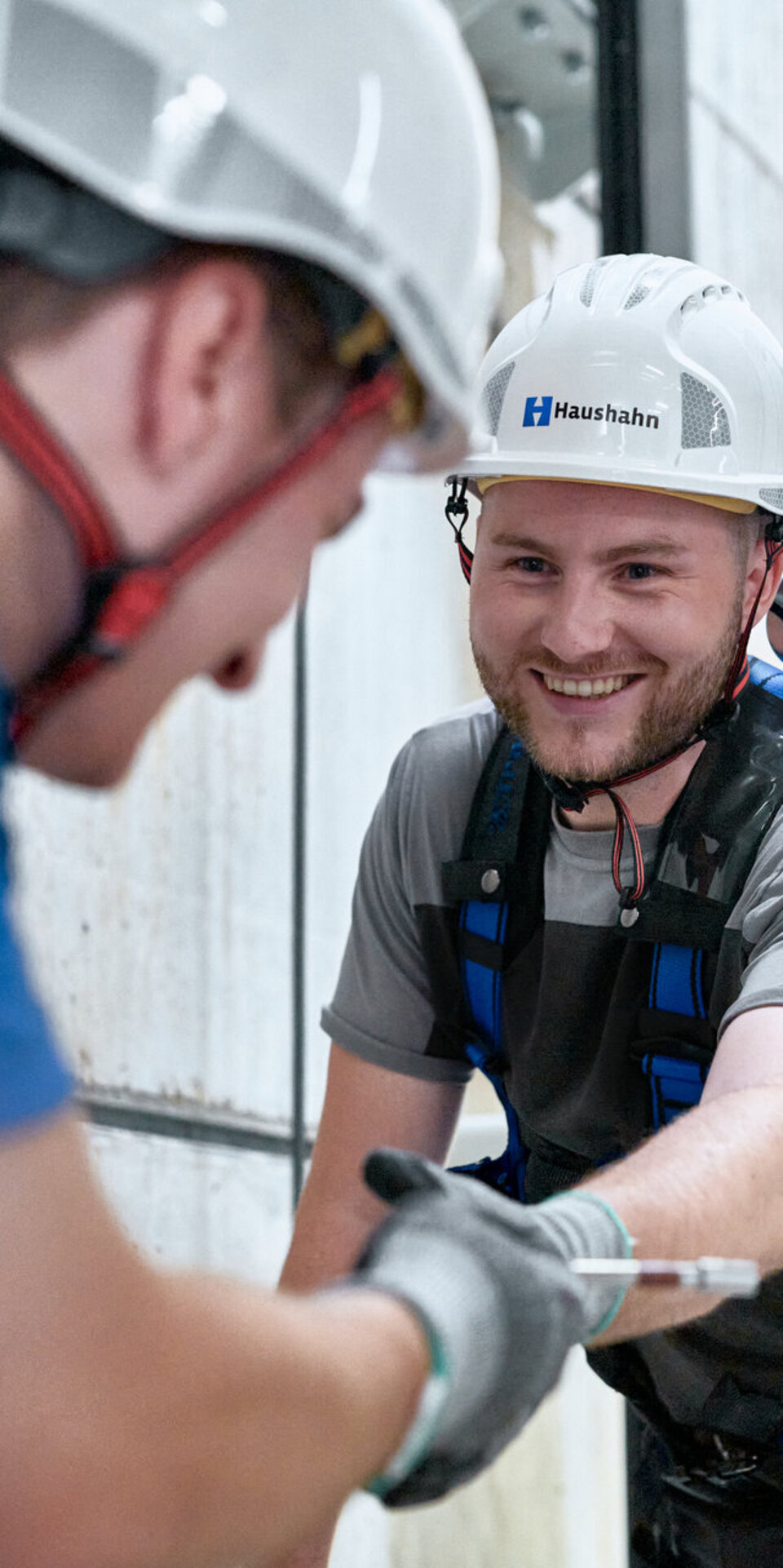 Zwei Arbeiter mit Schutzausrüstung und Helmen, einer mit der Aufschrift Haushahn, lächeln, während sie sich die Hand geben oder einen Gegenstand austauschen. Sie sind an Sicherheitsgurten in einer Industrieumgebung befestigt.