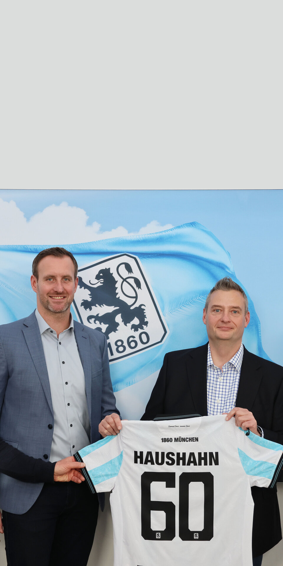 Vier Männer stehen in einer Halle, lächeln und halten ein Fußballtrikot mit dem Aufdruck HausHAHN 60 in der Hand. Hinter ihnen ist ein großes Bild einer blau-weißen Fahne mit dem Logo von 1860 München zu sehen.