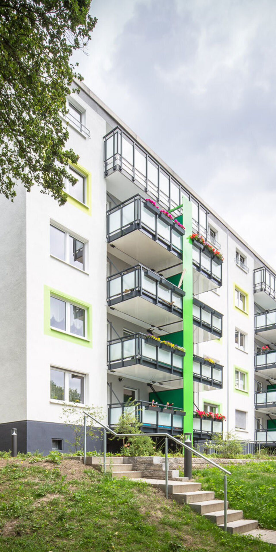 Ein modernes, weißes Wohnhaus mit grünen Akzenten und Balkonen, umgeben von Bäumen und einer Grünfläche. Eine Treppe führt zum Eingang hinauf. Der Himmel ist bewölkt.