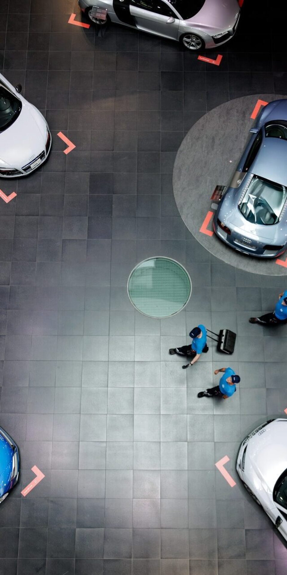 Draufsicht auf ein modernes Autohaus mit sechs Autos, die auf einem dunklen Kachelboden ausgestellt sind. Drei Personen in blauen Uniformen gehen in der Nähe der Mitte umher, und die Autos sind in kreisförmiger Anordnung angeordnet.