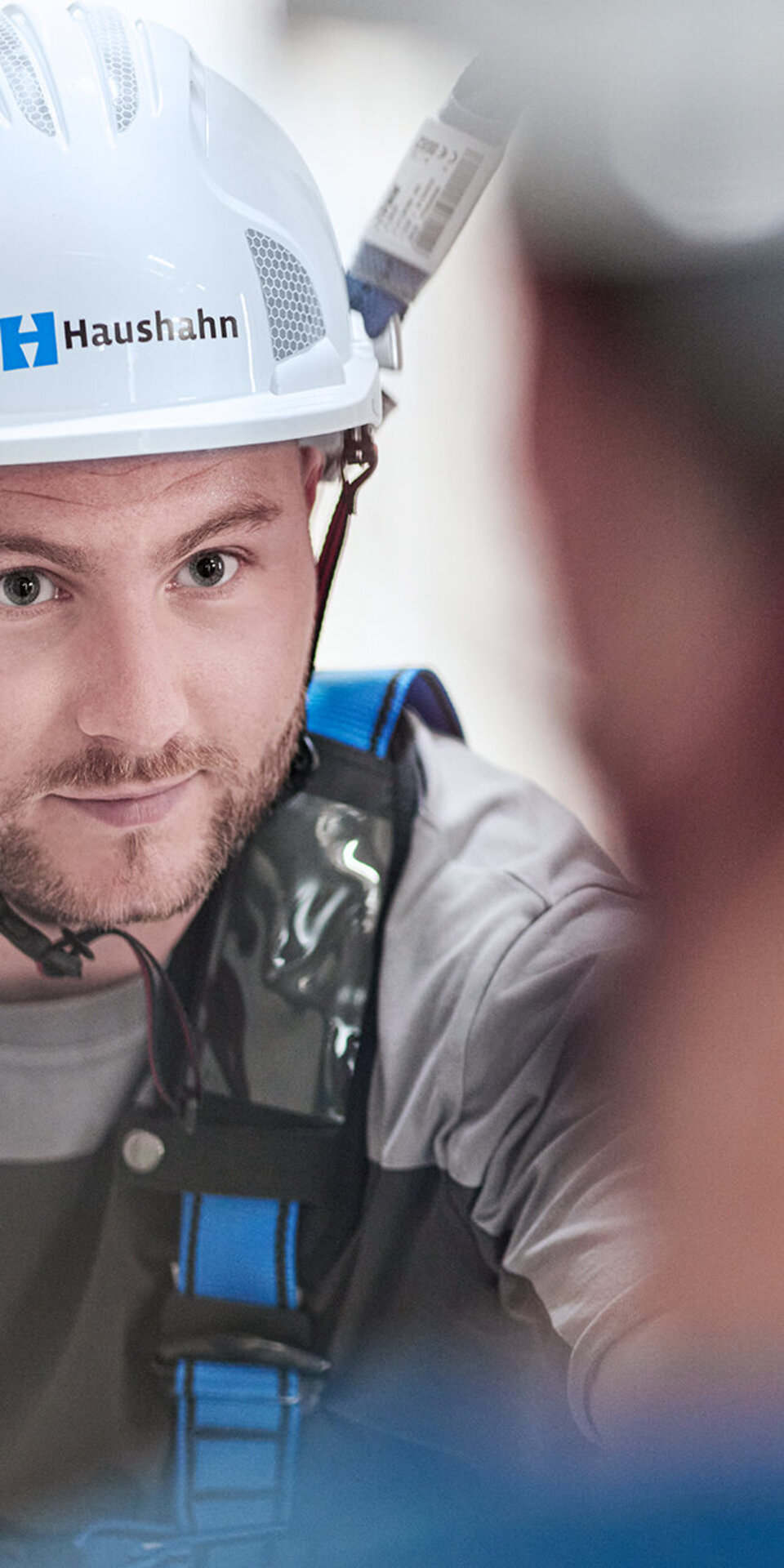Ein Mann, der einen weißen Schutzhelm mit der Aufschrift Haushahn und ein Sicherheitsgeschirr trägt, schaut aufmerksam auf eine unscharfe Person im Vordergrund.